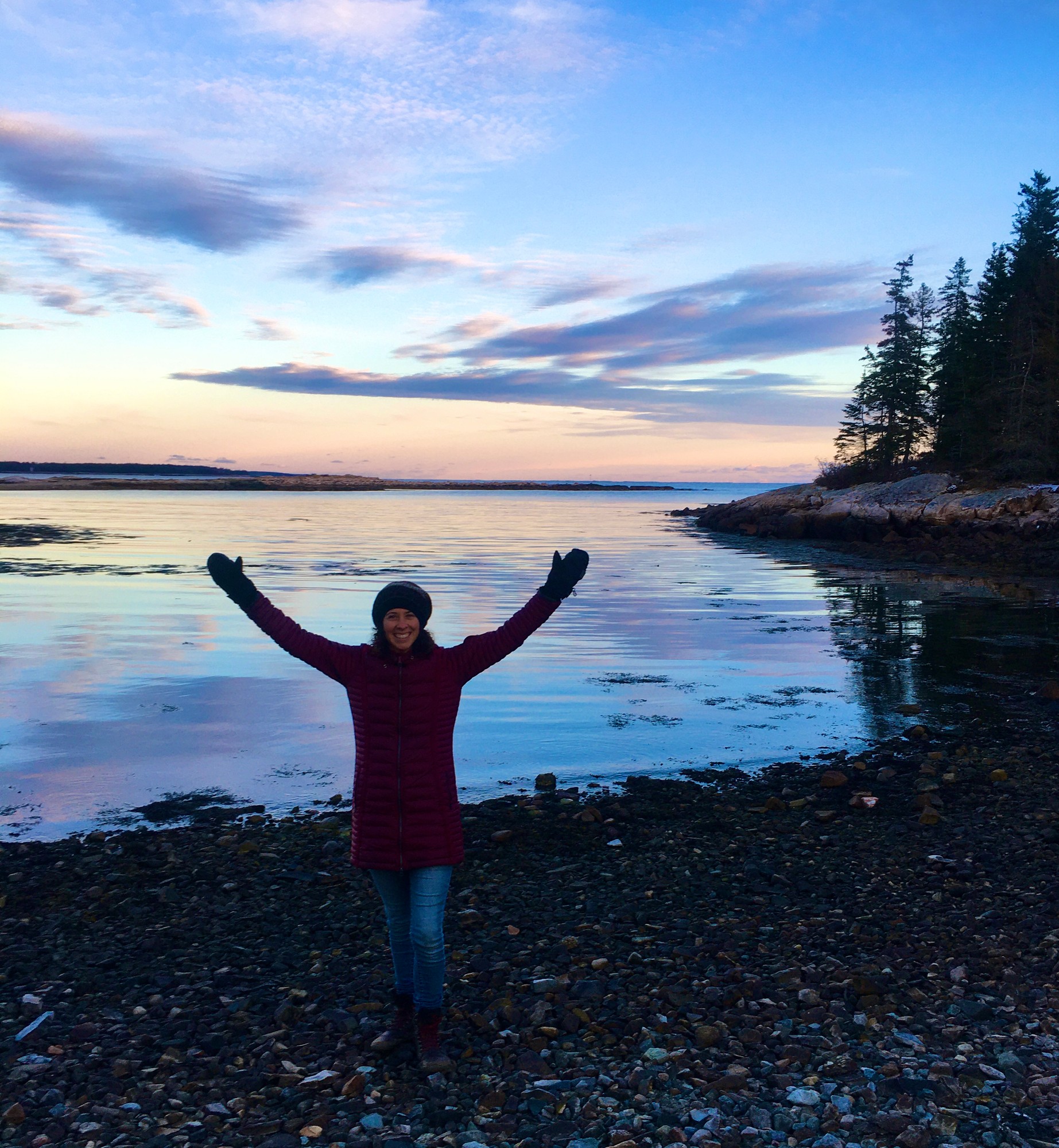 Vivian in a winter coat raising her arms on a pebbly shore at dusk