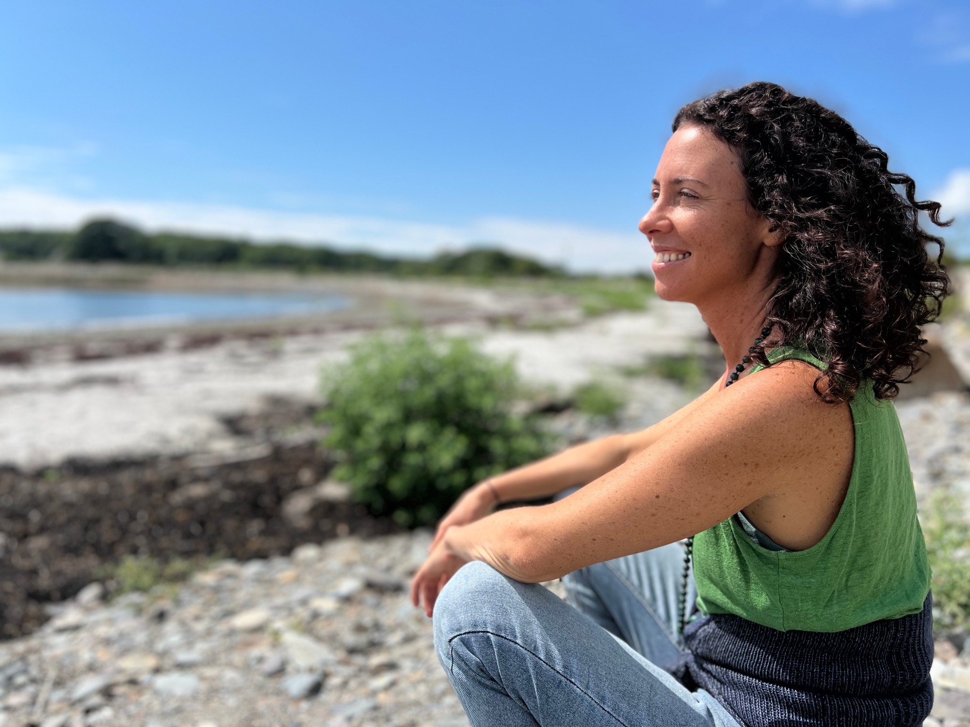 Vivian smiling, sitting on a pebbly shore under a blue sky
