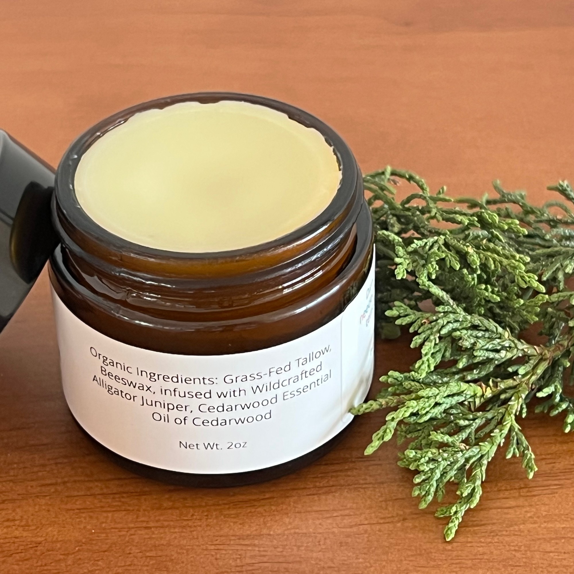 Open jar of Simmer Down Salve turned to its ingredients label, beside alligator juniper sprigs