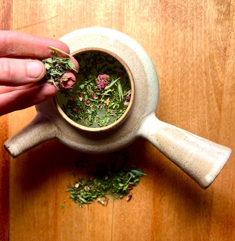 Vivian's hand adding dried herbs and rosebuds to a ceramic teapot