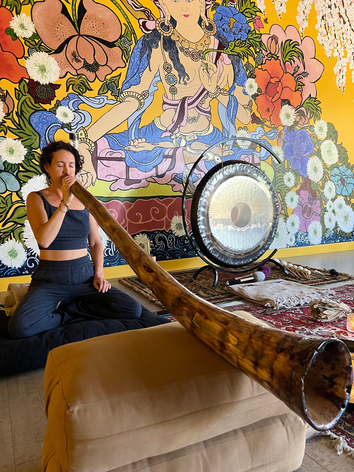 Vivian playing a long didgeridoo toward the camera, a gong and a painted mural behind her