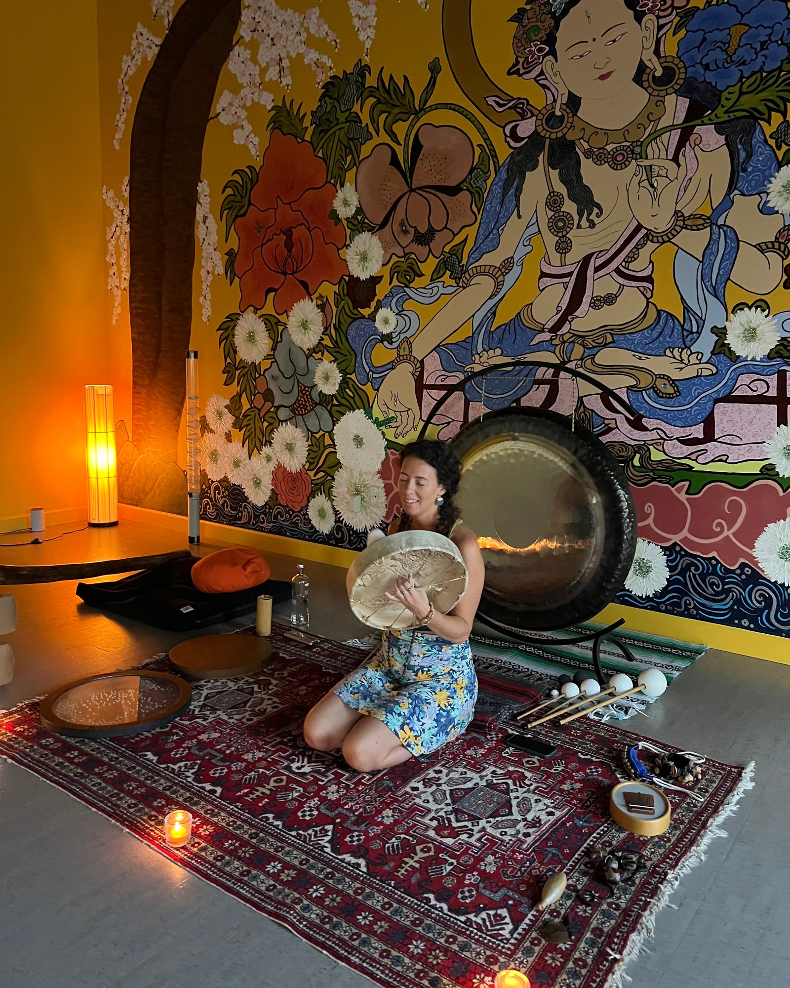 Vivian kneeling with a frame drum before a gong and a painted mural