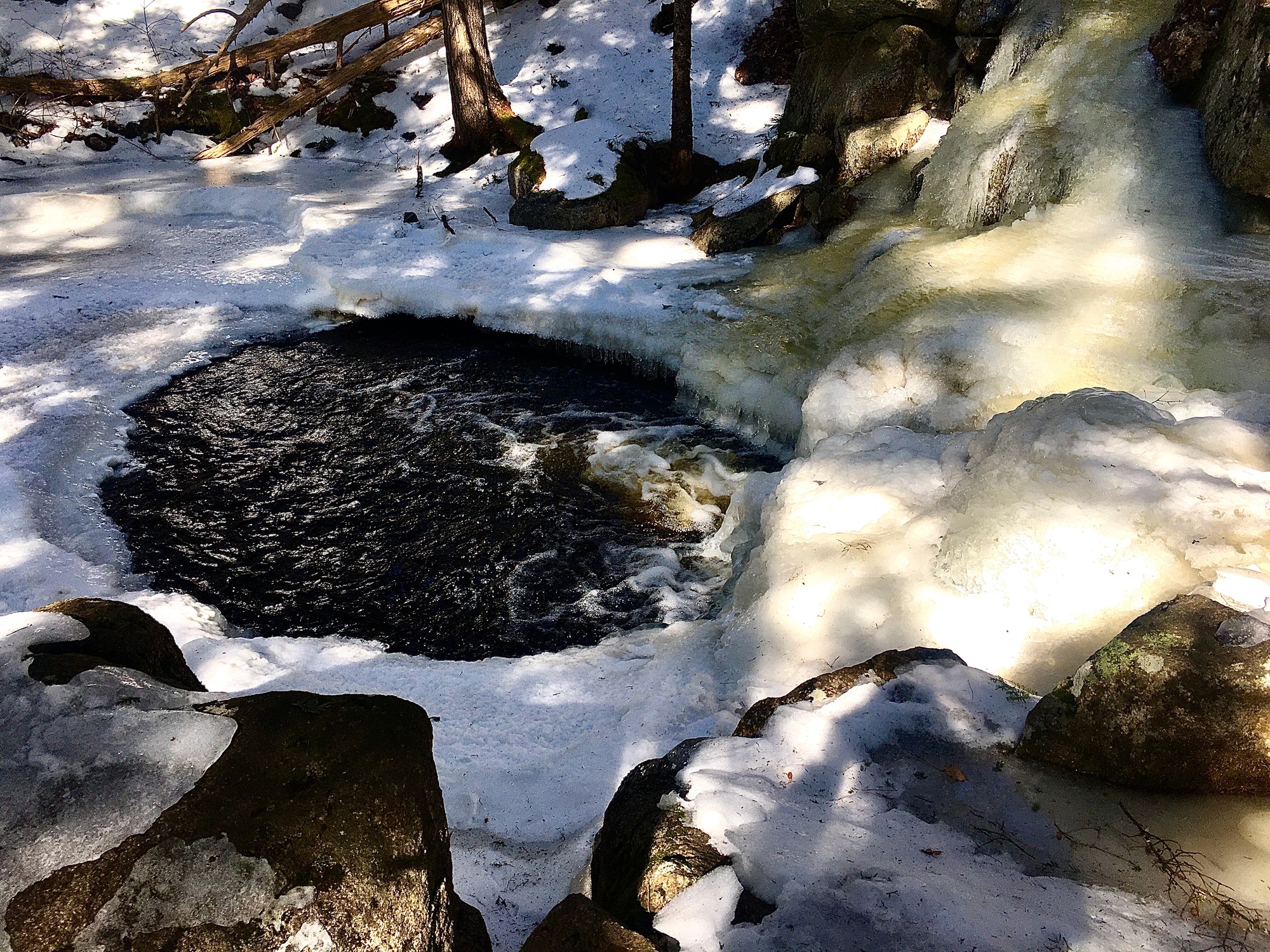 Dark pool ringed with ice below a partly frozen waterfall among snowy rocks